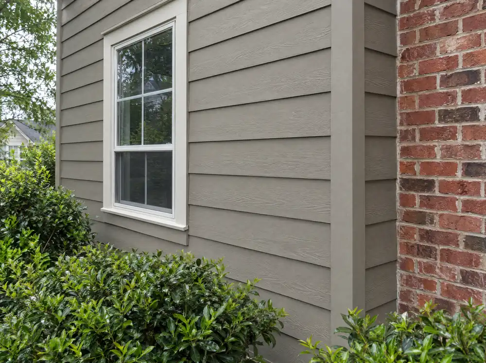 Warm gray lap siding meeting red brick, with a white window and shrubs